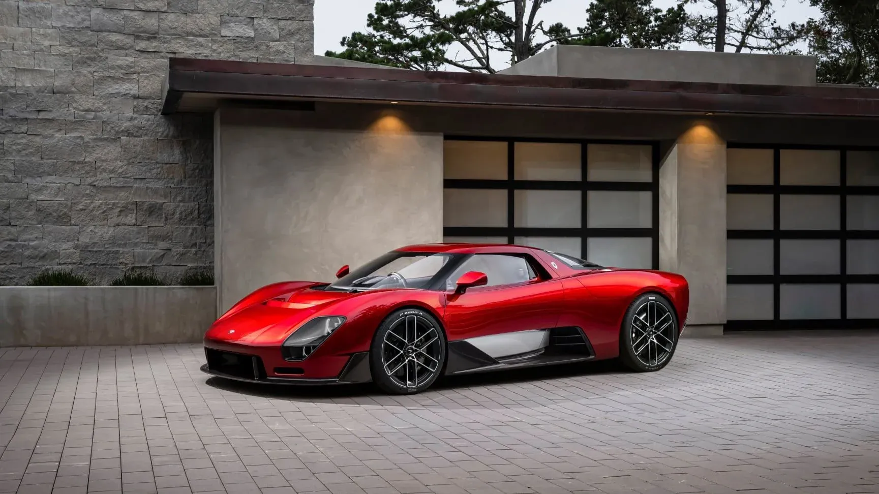 The McLaren McL 6GT concept in metallic red, seen from the front three-quarter on a paved forecourt in front of a stone and glass house at Monterey