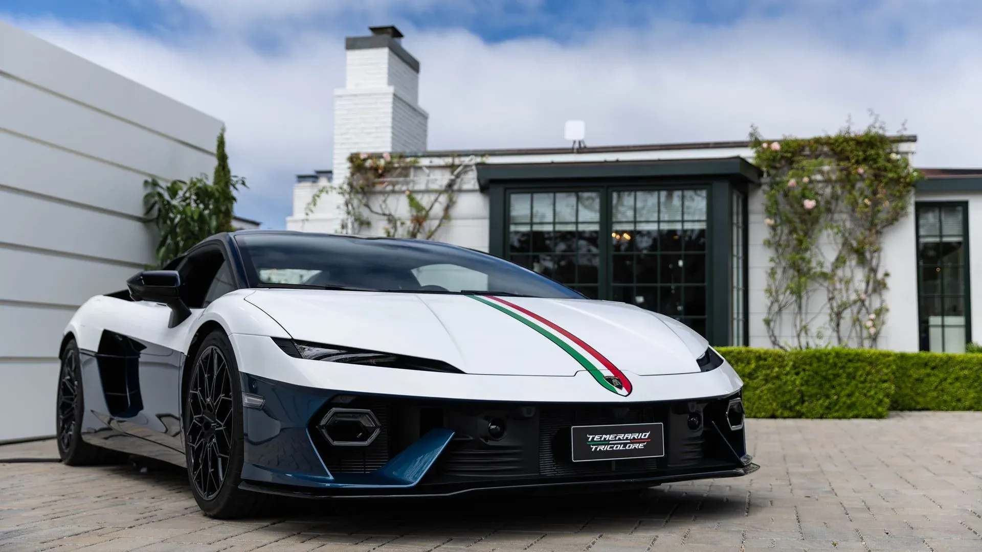 The Lamborghini Temerario Tricolore in white over blue with an Italian flag stripe on the bonnet, seen from the front three-quarter on a paved forecourt at Monterey