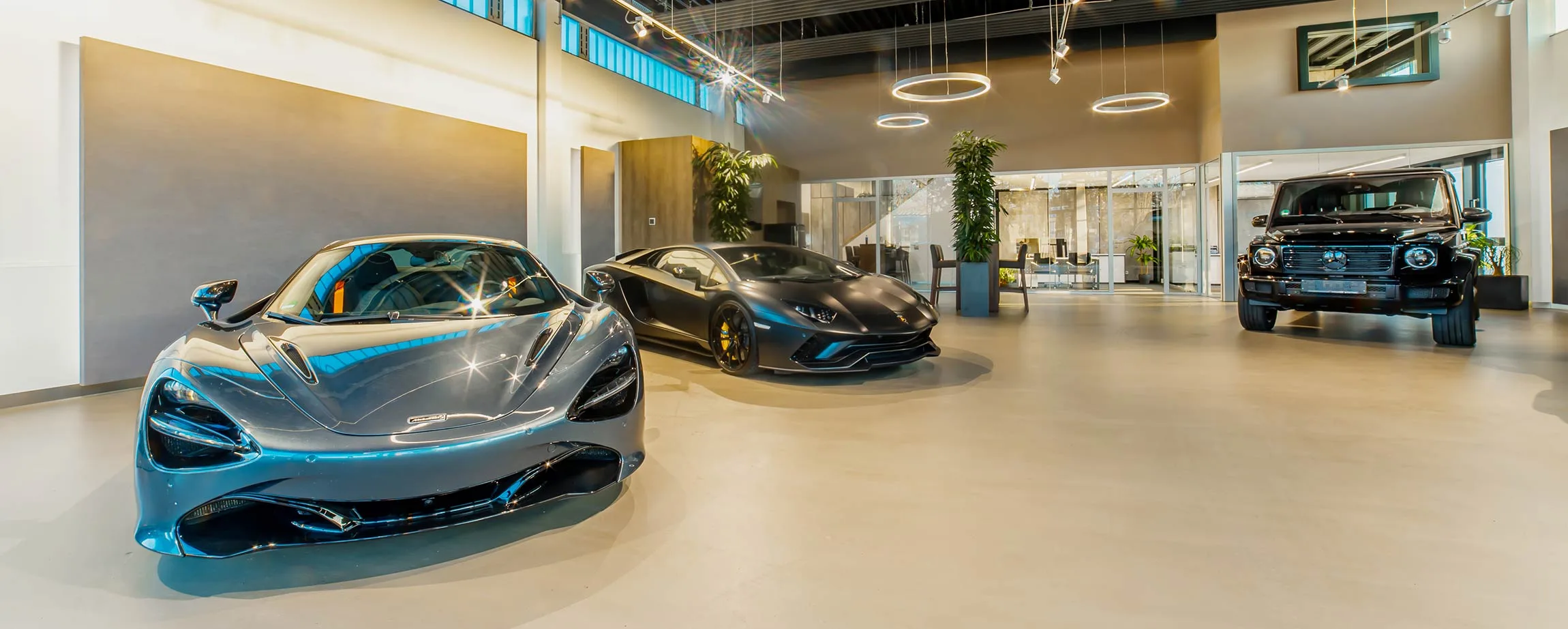Le showroom d'Akpinar-Automobile à Holzgerlingen, une McLaren 720S grise et une Lamborghini Aventador S noir mat au premier plan, un Mercedes-Benz Classe G noir devant les bureaux vitrés