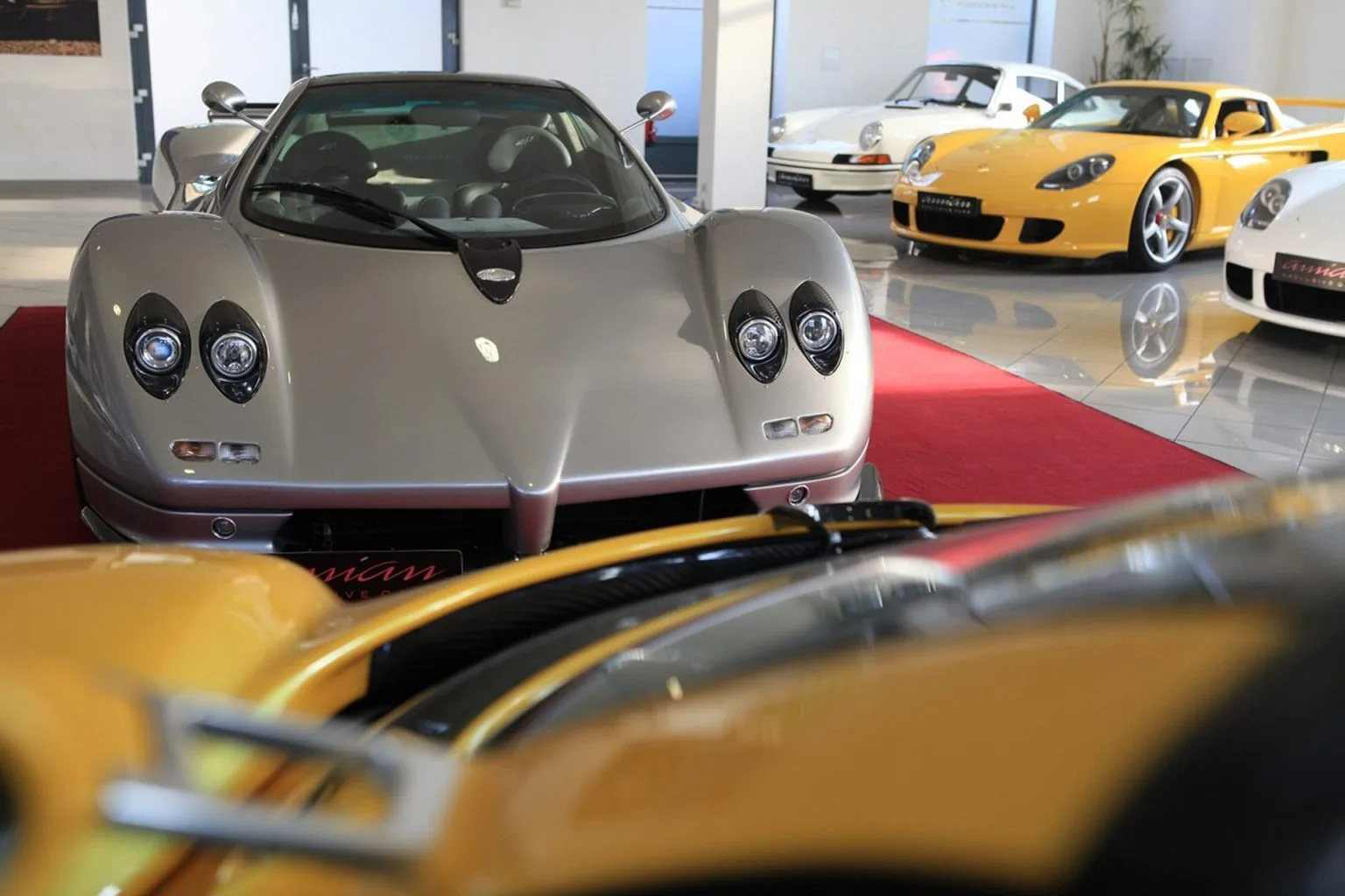 Une Pagani Zonda S argentée vue de face dans le showroom d'Amian Exclusive Cars, avec une Porsche Carrera GT jaune et deux voitures de sport blanches derrière elle