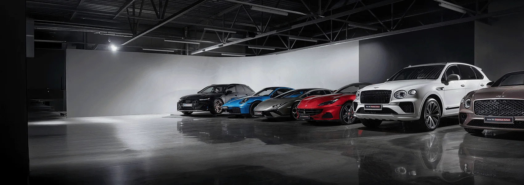 Die Halle von Auto 100 Premium Select in Tallinn, eine Reihe von Fahrzeugen unter Industriebeleuchtung: ein schwarzer Audi Kombi, ein blauer Porsche 911, ein grauer Lamborghini Huracán, ein roter Ferrari, ein weißer Bentley Bentayga und ein bronzefarbener Bentley
