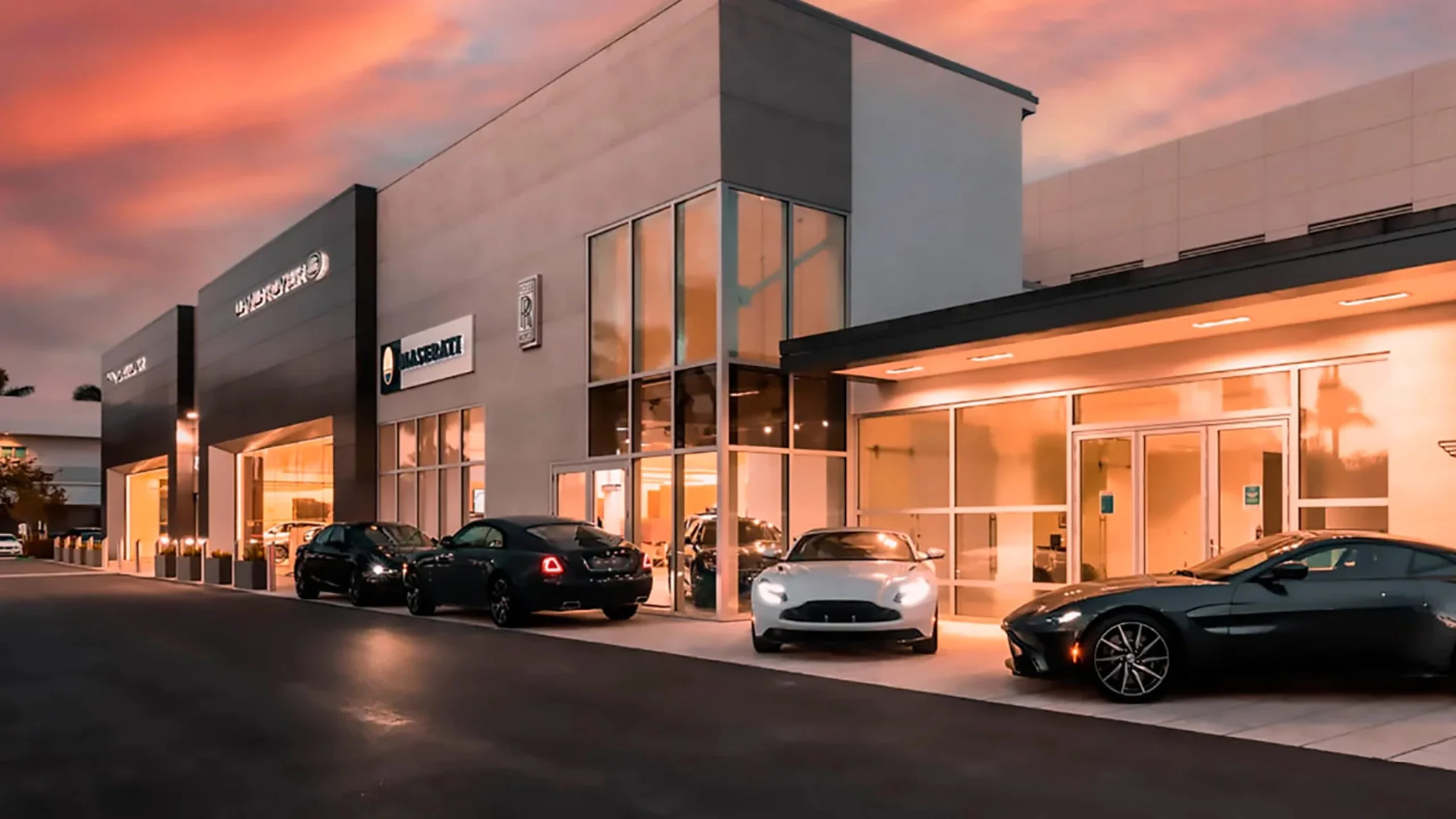 Un showroom automobile moderne à façade vitrée éclairée au crépuscule sous un ciel rose, une Aston Martin noire et une grand tourisme blanche garées devant, en couverture d'Autocenter Sportive
