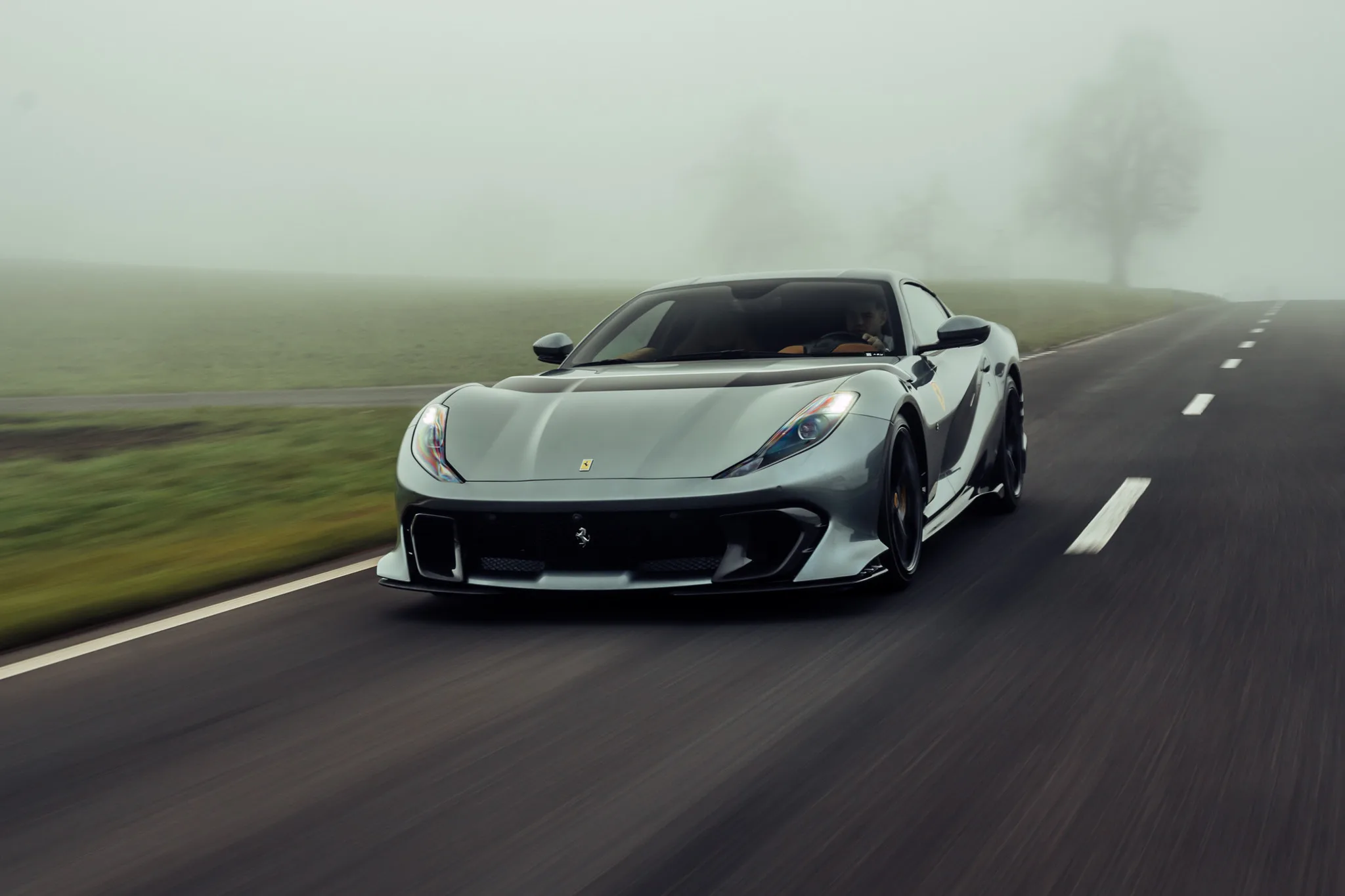 Une Ferrari 812 Competizione grise roulant vers l'objectif sur une route de campagne dans la brume, cuir fauve visible derrière le pare-brise, talus vert sur la gauche