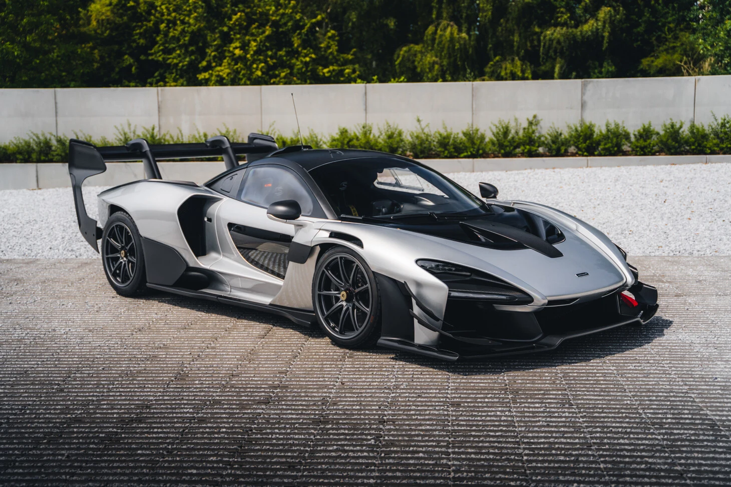 A McLaren Senna GTR in silver and exposed carbon, parked on the paved forecourt of Bavaria Motors in Harelbeke, against the concrete wall and clipped hedge the dealer photographs its cars in front of