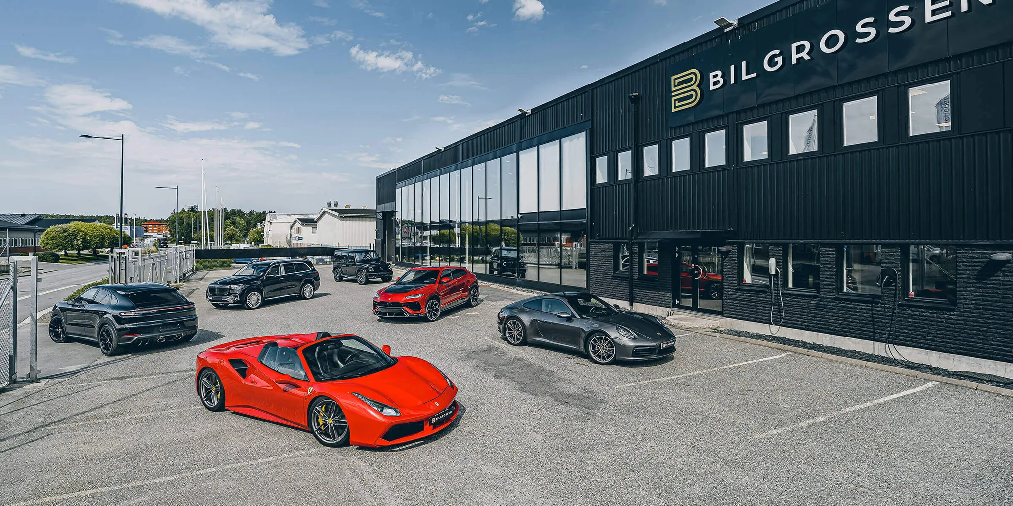 Le bâtiment de Bilgrossen à Täby, façade noire et logo éclairé, une Ferrari 488 Spider rouge au premier plan, une Porsche 911, un Lamborghini Urus, un Mercedes-Maybach GLS, un Mercedes Classe G et un Porsche Cayenne sur le parvis