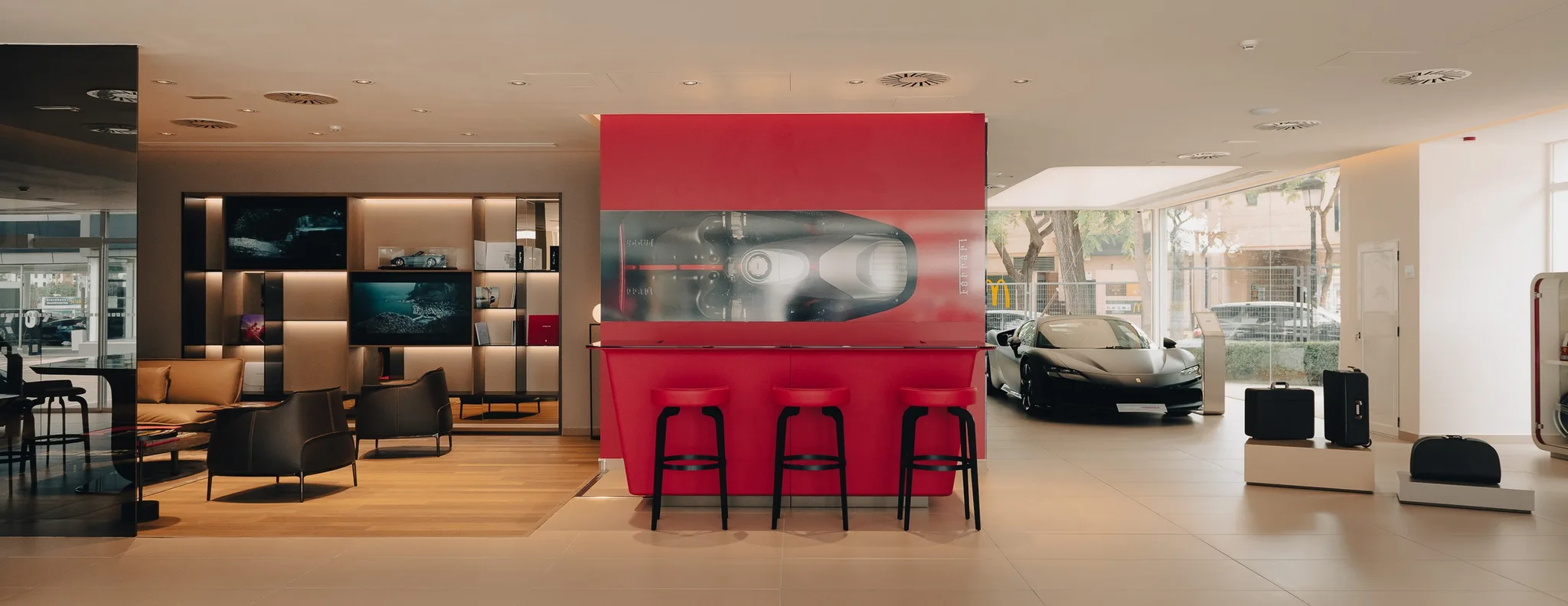 Der Showroom von C. de Salamanca in San Pedro Alcántara, mit rotem Tresen und drei Hockern gegenüber einer Lounge aus Ledersesseln, ein schwarzer Ferrari an der verglasten Front