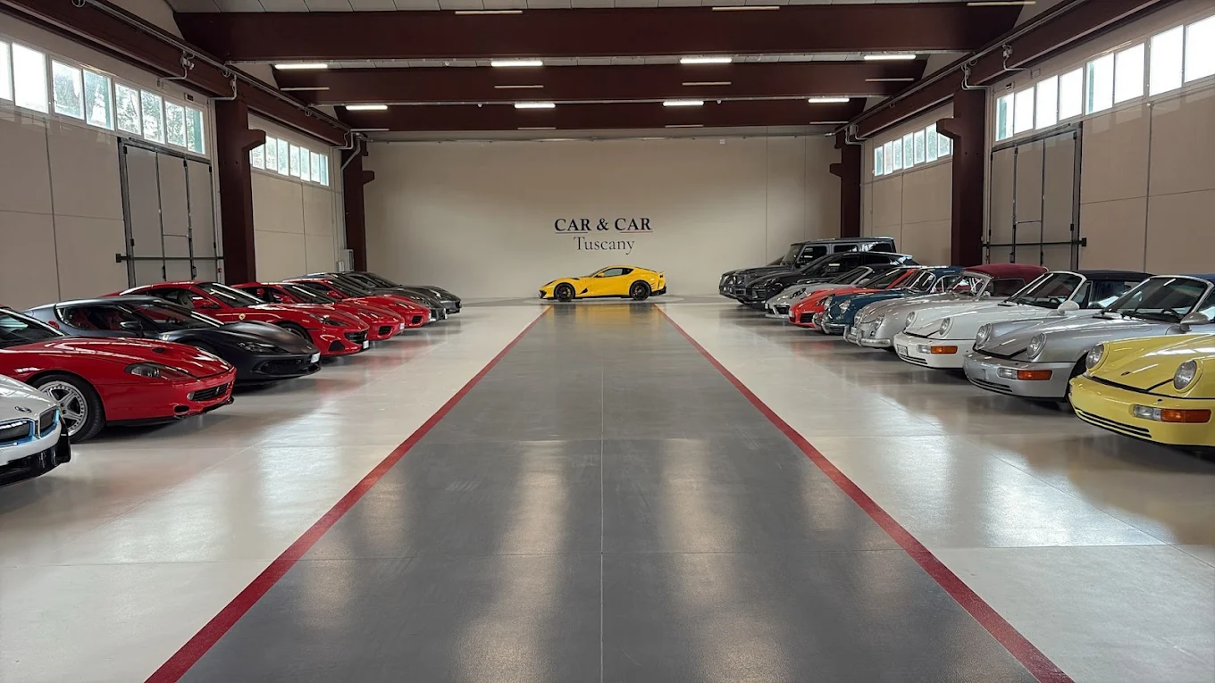Der Ausstellungsraum von Car & Car Tuscany der Länge nach gesehen, links Ferrari und rechts luftgekühlte Porsche beiderseits eines rot gefassten Mittelgangs, ein gelber Ferrari 812 allein am hinteren Ende unter dem an die Wand gemalten Schriftzug