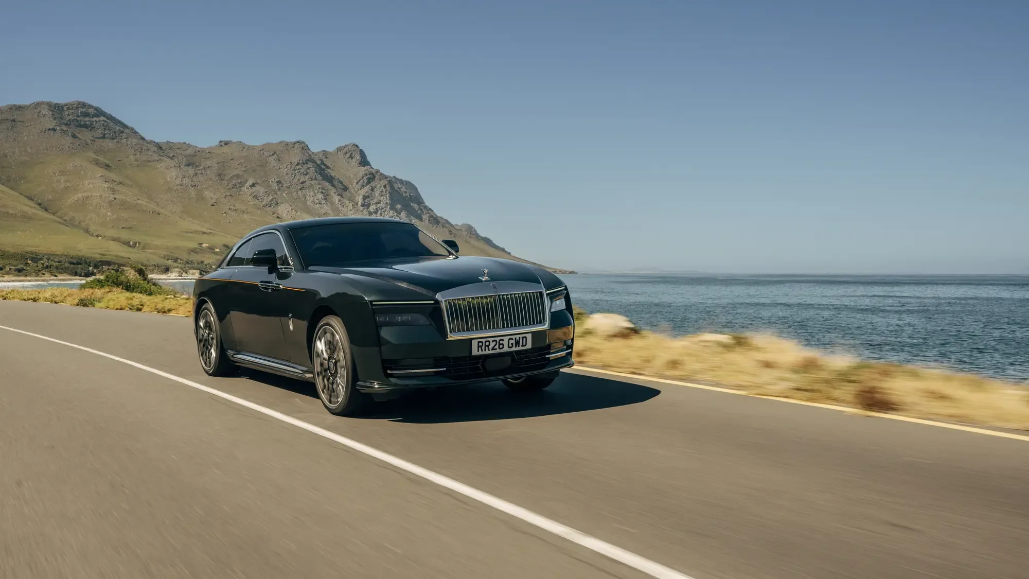 Une Rolls-Royce Spectre vert foncé sur une route côtière, la mer à sa droite et des collines verdoyantes derrière, sous un ciel dégagé