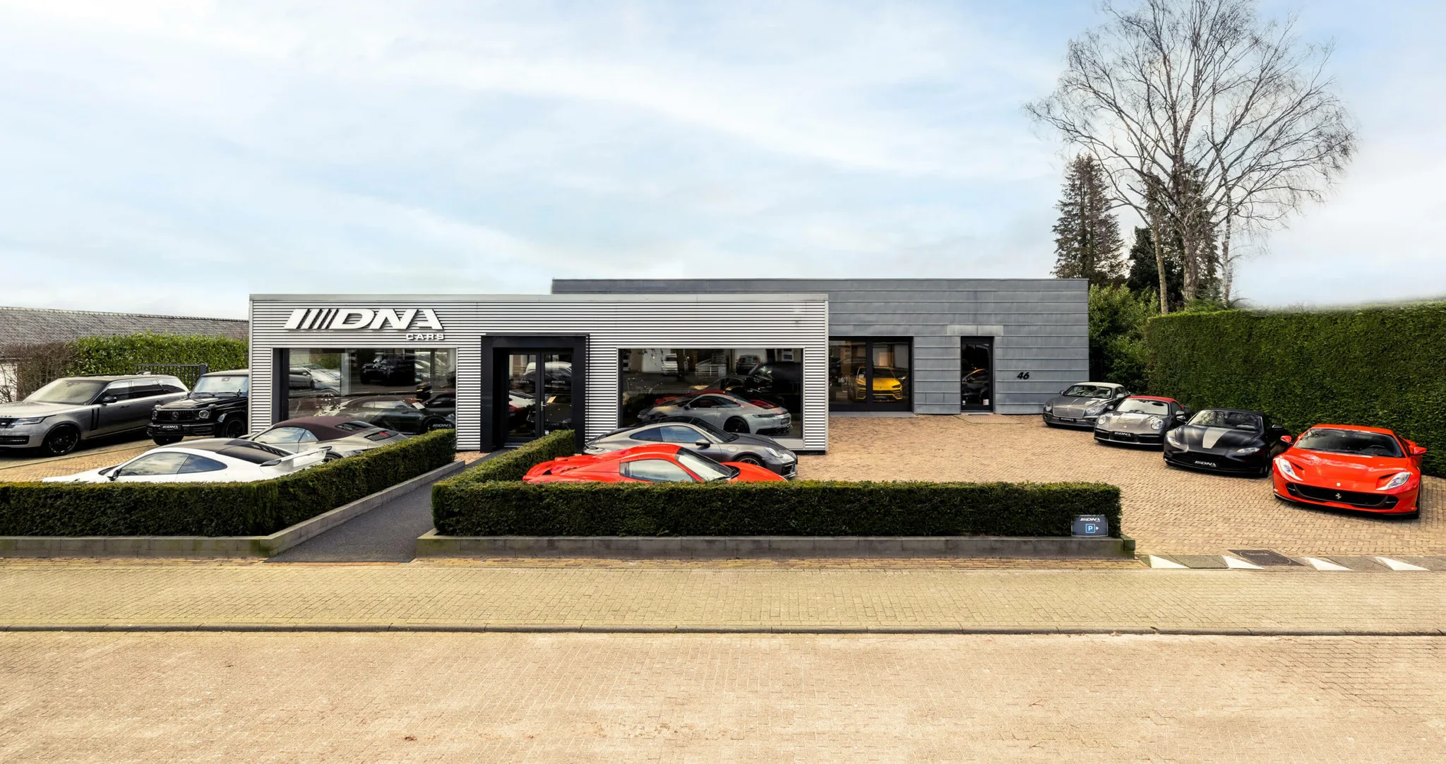 The DNA Cars showroom in Naarden, a long grey building with the DNA logo, cars parked in front including red Ferraris, under a spring sky