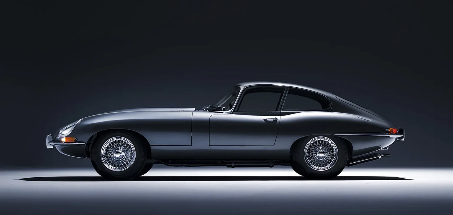 A silver-grey Jaguar E-Type Series 1 fixed-head coupé in pure profile, wire wheels and chrome bumpers, lit in a dark studio