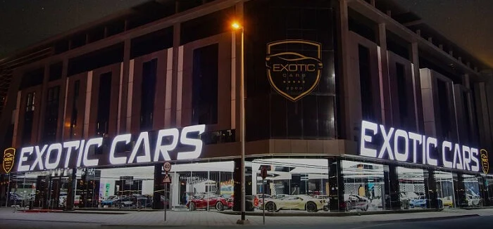 The Exotic Cars Dubai showroom at night on Sheikh Zayed Road: the illuminated EXOTIC CARS lettering runs along two facades, the crest with the year 2003 sits above the corner, and supercars are lined up behind the glass on the ground floor