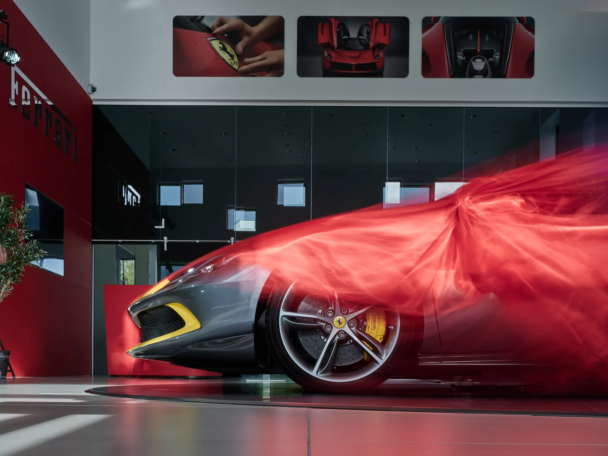 Une Ferrari sous une housse de soie rouge sur le plateau tournant du showroom Formula Automobile à Kgs. Lyngby, son bouclier avant souligné de jaune et son splitter en carbone laissés découverts, le mot Ferrari découpé dans le mur rouge à gauche et trois photographies rétroéclairées au-dessus de la mezzanine vitrée