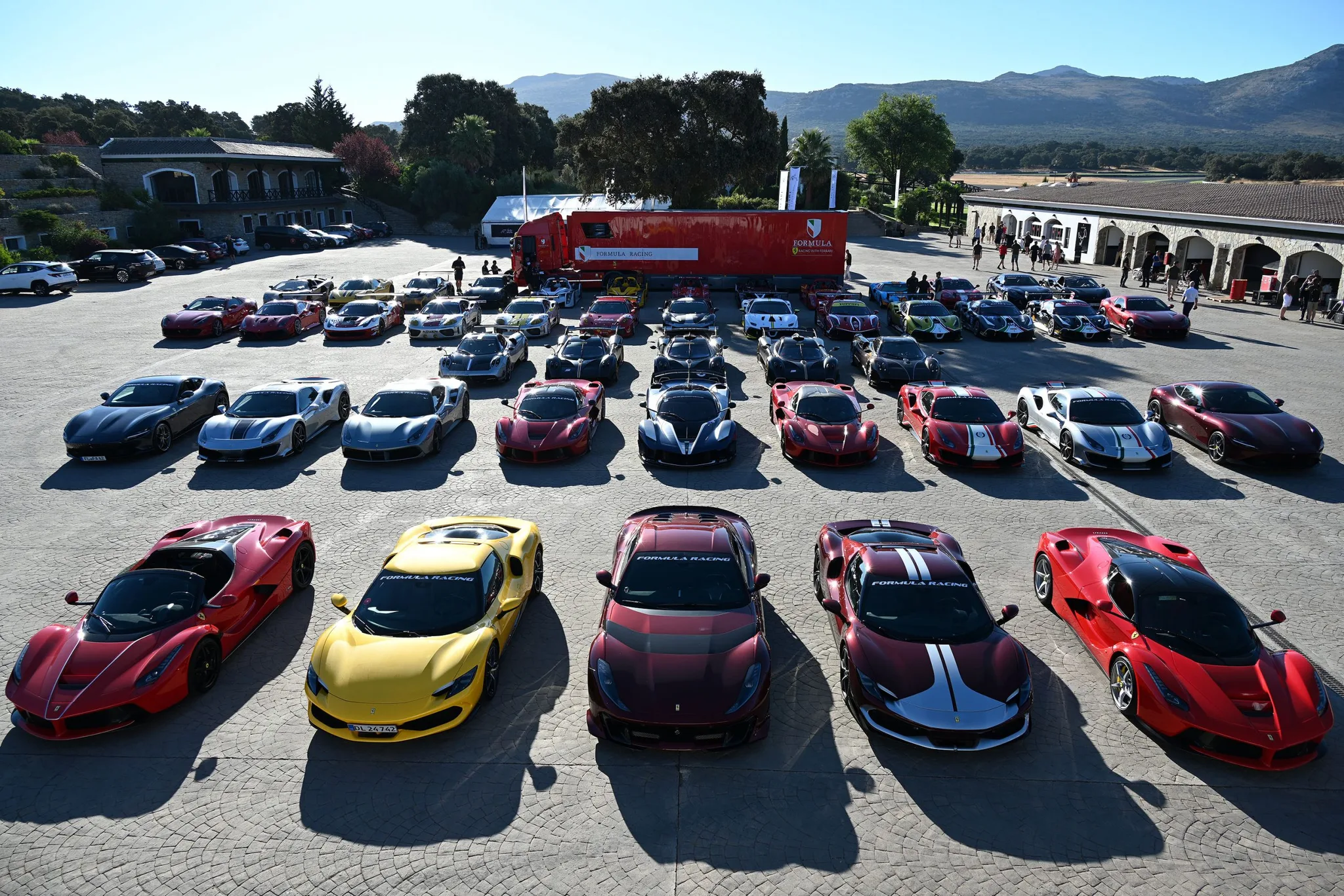 Une quarantaine de Ferrari et de Pagani alignées en rangs sur une cour ensoleillée lors d'un rassemblement Formula Racing, cinq LaFerrari et 296 GTB au premier rang, le semi-remorque rouge de Formula Racing garé derrière et des collines boisées à l'horizon