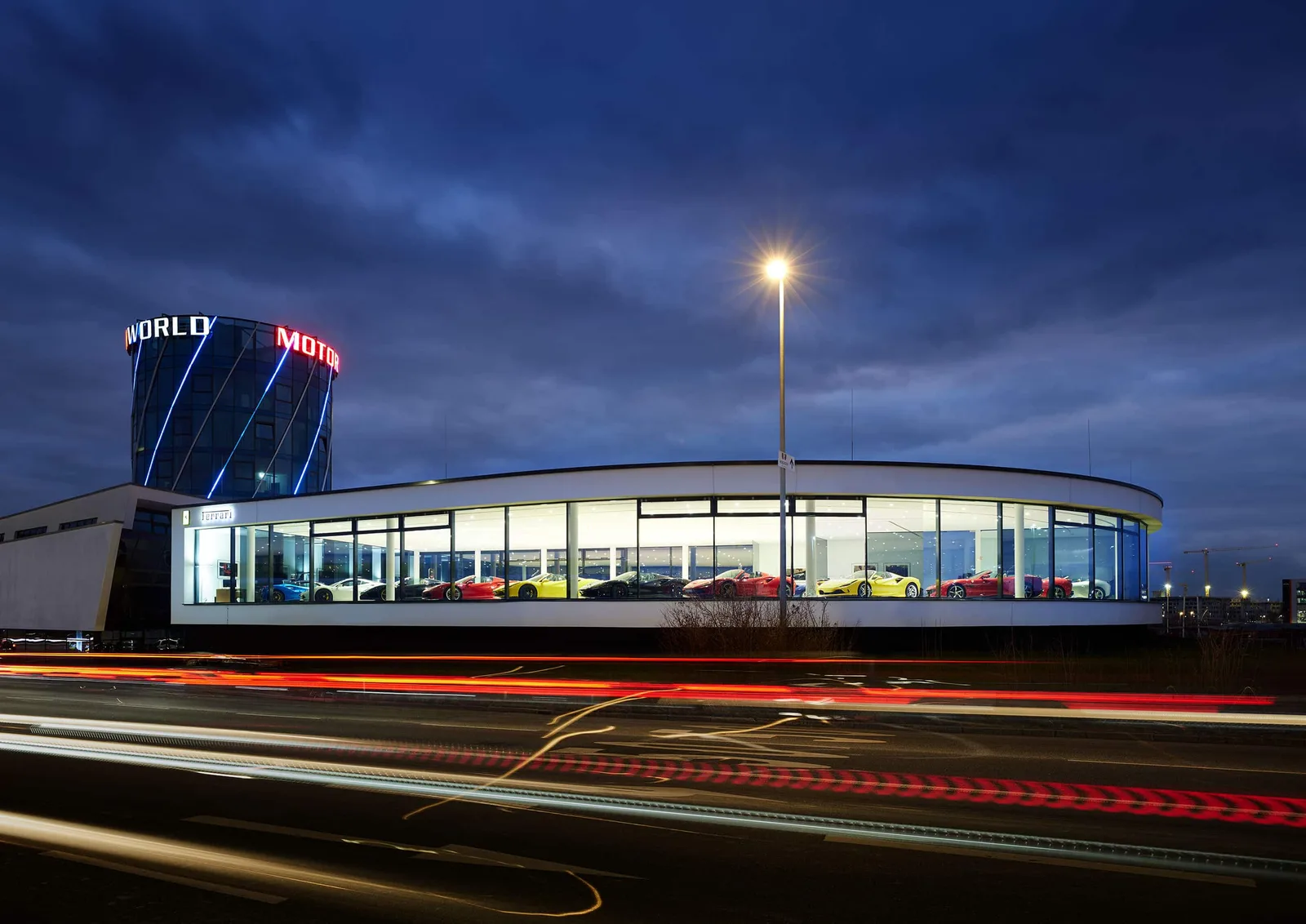 The Gohm Böblingen showroom at dusk, its curved glass front lit from within and lined with Ferraris, beneath the Motorworld tower