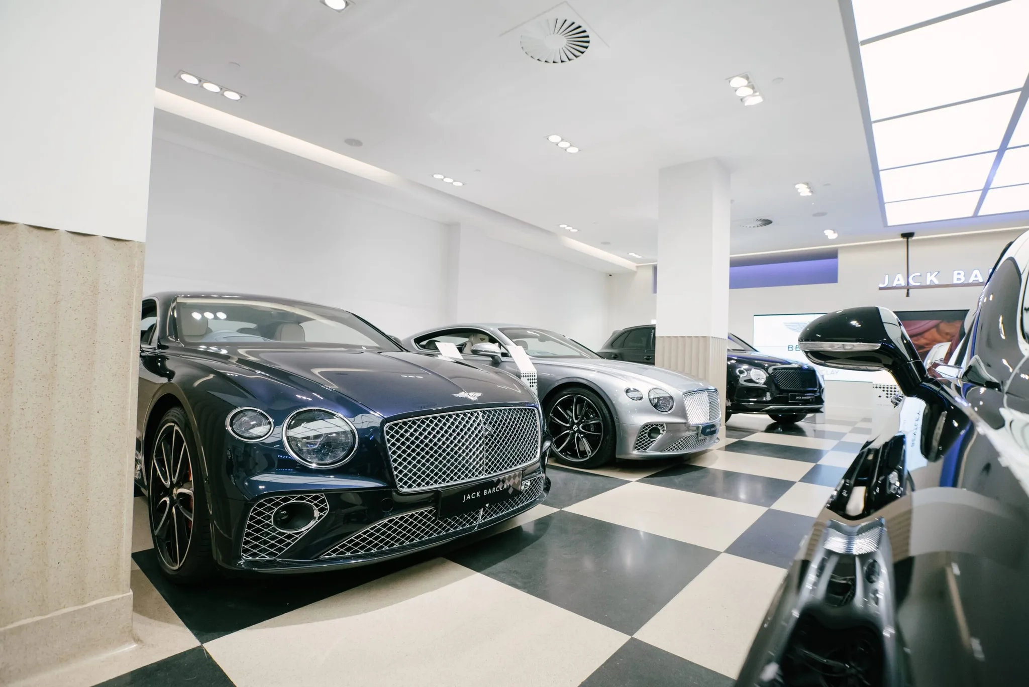 Ein dunkelblauer Bentley Continental GT auf dem schwarz-weiß karierten Boden des Showrooms von Jack Barclay Bentley am Berkeley Square, dahinter ein silberner Continental GT und ein Bentayga unter einem Glasdach