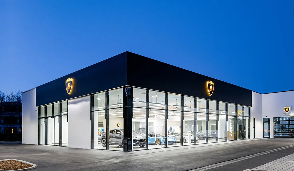 The Lamborghini München building on the Leonrodstraße at dusk, its glass showroom lit with an Urus and a Huracán inside, illuminated Lamborghini shields on the black façade