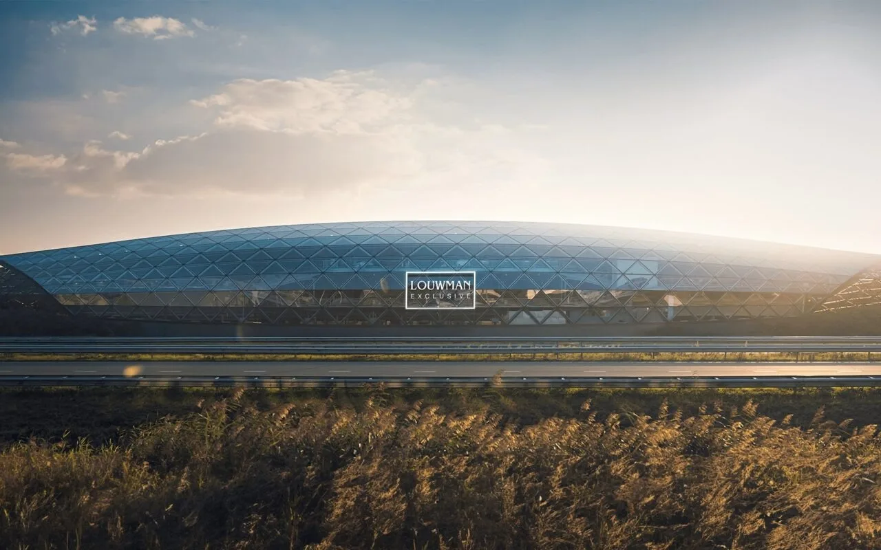 Le showroom Louwman Exclusive d'Utrecht au soleil couchant, un long bâtiment de verre sous une toiture réticulée courbe, vu depuis l'autoroute qui longe le site