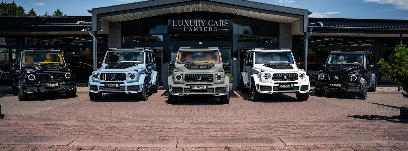 Cinq Mercedes Classe G transformés par Brabus alignés devant le showroom de Luxury Cars Hamburg, en noir, bleu clair, gris, blanc et noir, sous le lettrage de la façade sombre à pignon