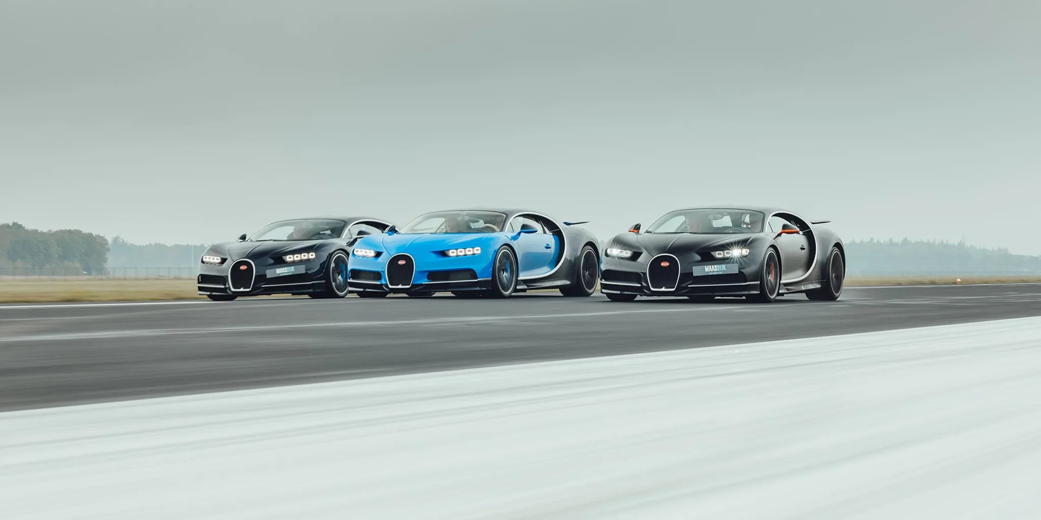 Trois Bugatti Chiron roulant de front sur une piste sous un ciel gris, une bleue et noire entre deux sombres, chacune portant une plaque Maasdijk Automotive