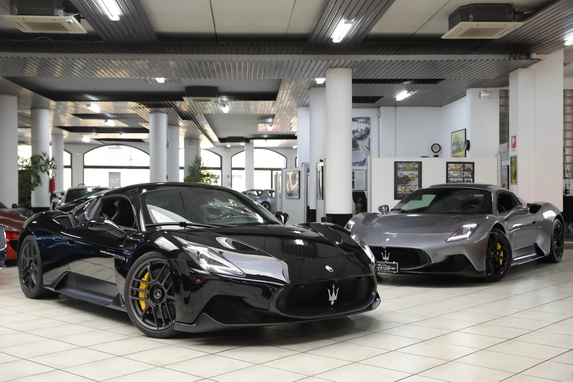 Deux Maserati MC20, une noire et une grise, garées côte à côte sous le plafond voûté du showroom MC Motors à Milan