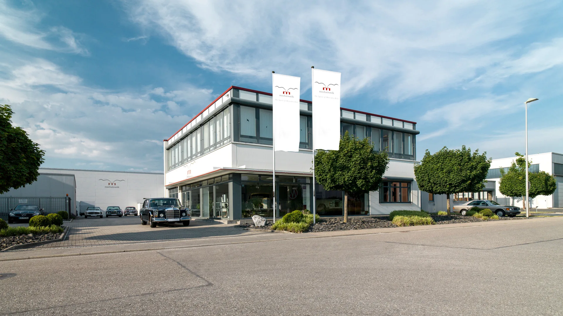 Le siège de Mechatronik à Pleidelsheim, au nord de Stuttgart : un bâtiment de deux niveaux à façade vitrée et corniche rouge, deux drapeaux blancs au logo de la maison, une Mercedes-Benz 600 noire et une rangée de Mercedes classiques sur l'avant-cour