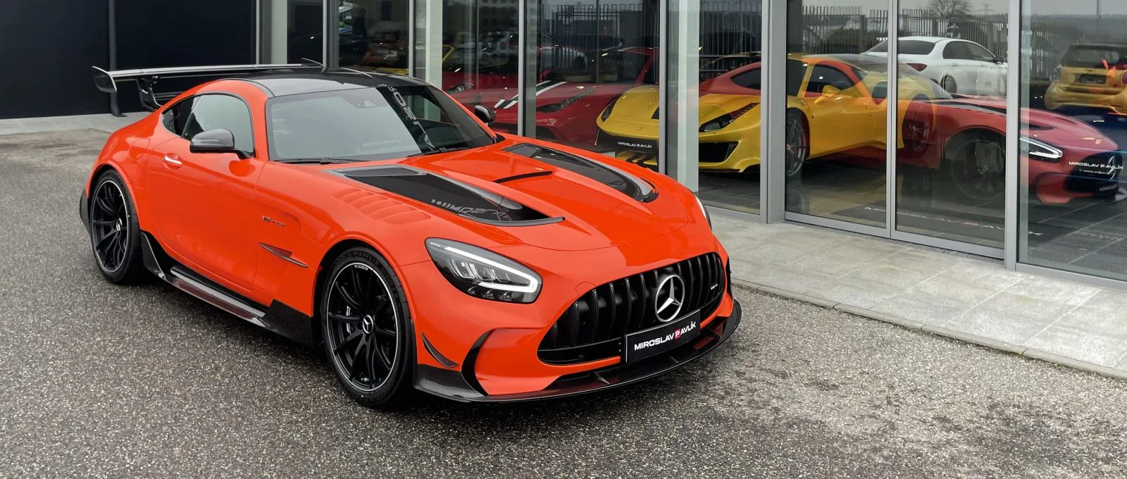 Ein orangefarbener Mercedes-AMG GT Black Series vor dem Showroom von Miroslav Pavlík, dahinter durch das Glas Sportwagen von Ferrari und Porsche