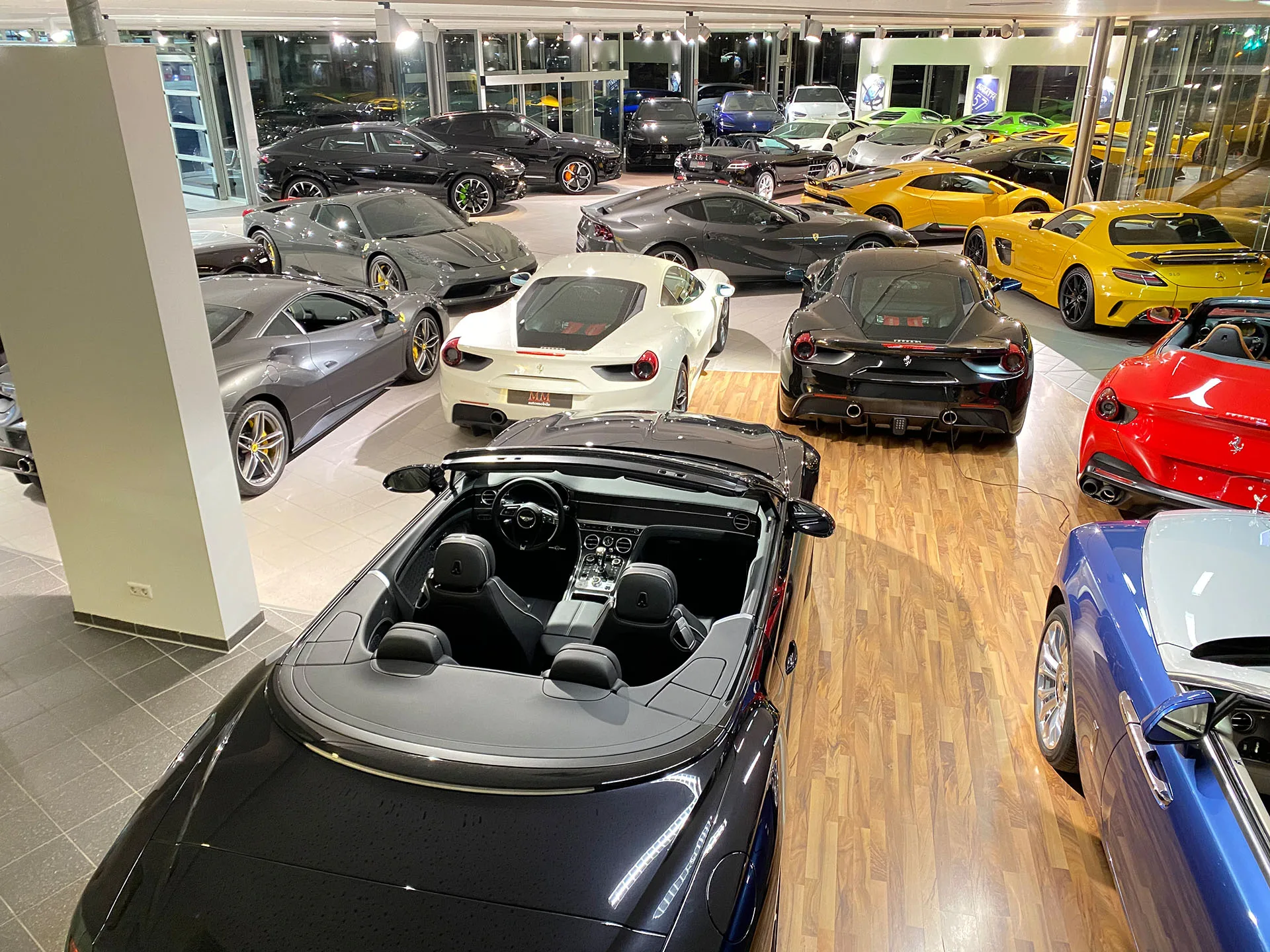 Le showroom MM Automobile à Hanovre vu de haut, une douzaine de voitures de sport rangées côte à côte sur un sol de bois et de carrelage, dont une Ferrari 488 blanche, une Ferrari cabriolet rouge et une Bentley Continental GT décapotable sombre au premier plan