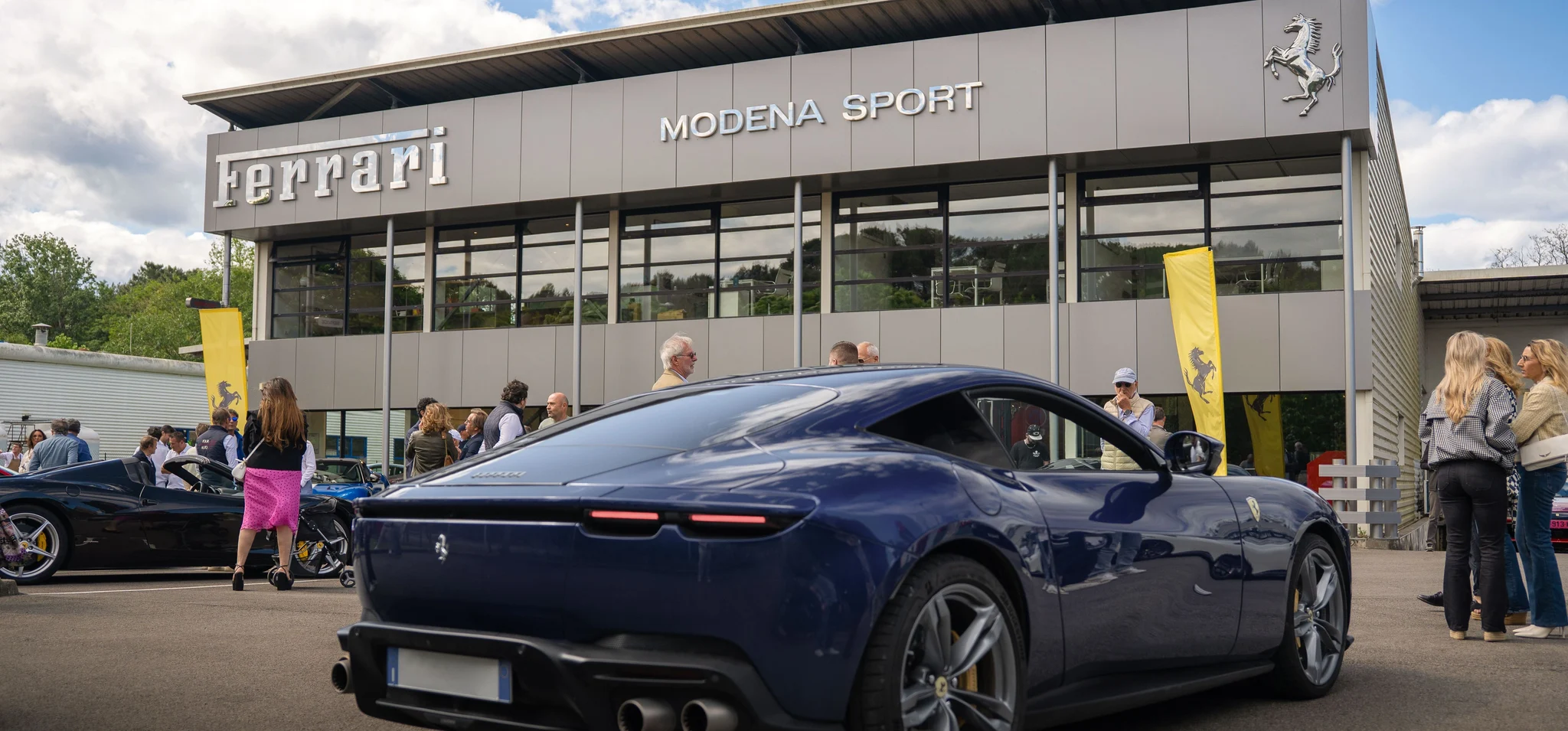 Le showroom Modena Sport de Biarritz lors d'une journée portes ouvertes, avec le lettrage Ferrari, l'inscription MODENA SPORT et le cheval cabré sur sa façade grise, une Ferrari Roma bleu foncé garée devant et des visiteurs entre des oriflammes Ferrari jaunes