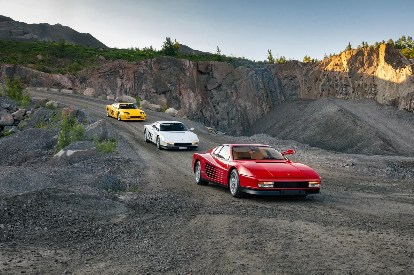 Three Ferraris on a gravel road through a Swedish quarry at golden hour, a red Testarossa in the foreground, a white Testarossa and a yellow F355 behind it, the cut rock face and spoil heaps rising on either side