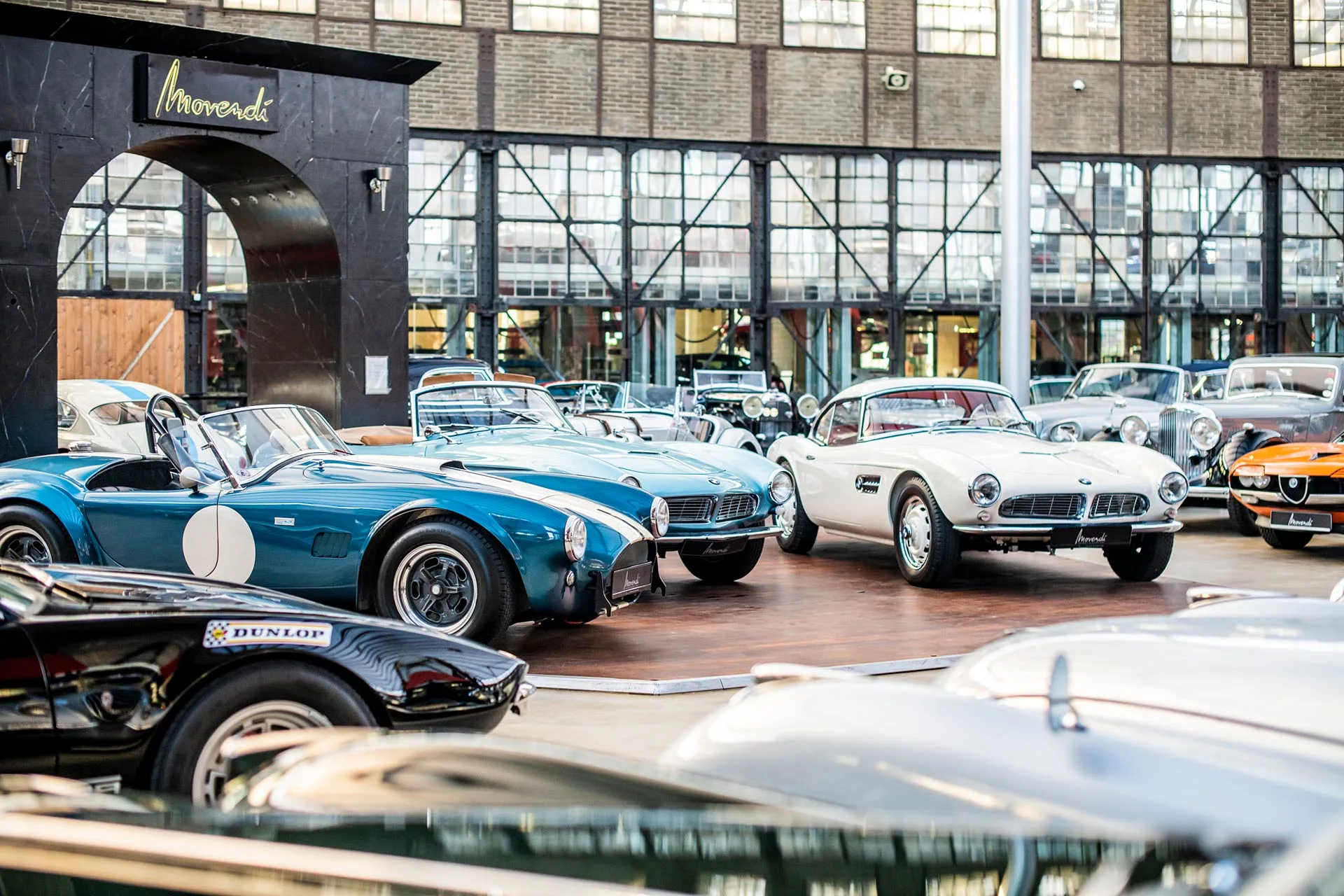 Klassische Fahrzeuge, darunter zwei AC Cobra und ein weißer BMW 507, unter dem Movendi-Schriftzug in der Classic Remise Düsseldorf