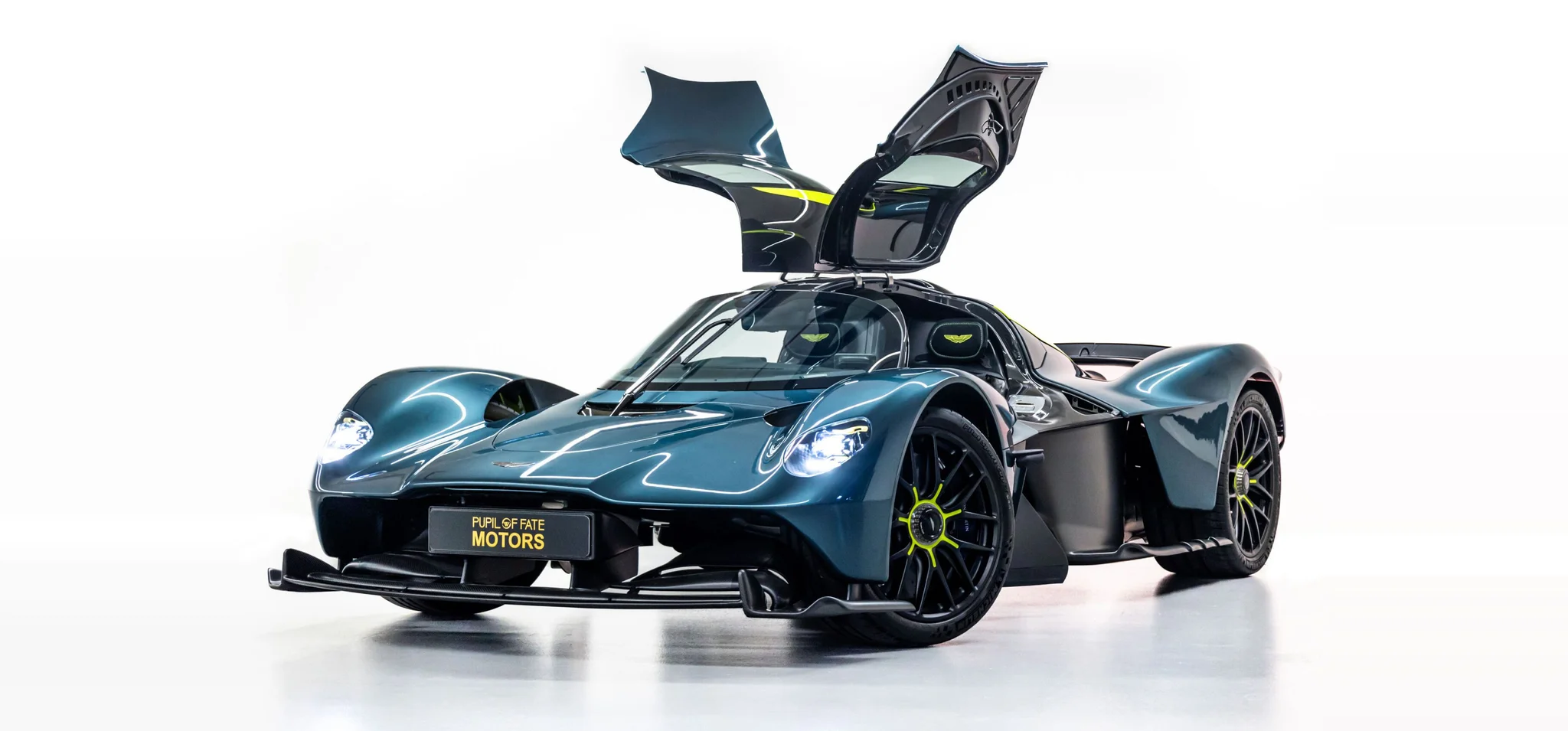 Une Aston Martin Valkyrie bleu pétrole foncé, les deux portes papillon levées et les moyeux vert citron, photographiée dans un studio blanc avec une plaque Pupil of Fate Motors à l'avant