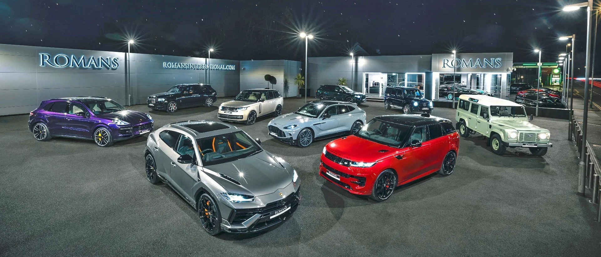 Le site Romans International à Banstead de nuit, les deux bâtiments du showroom éclairés et des supercars et SUV de luxe alignés sur le parvis