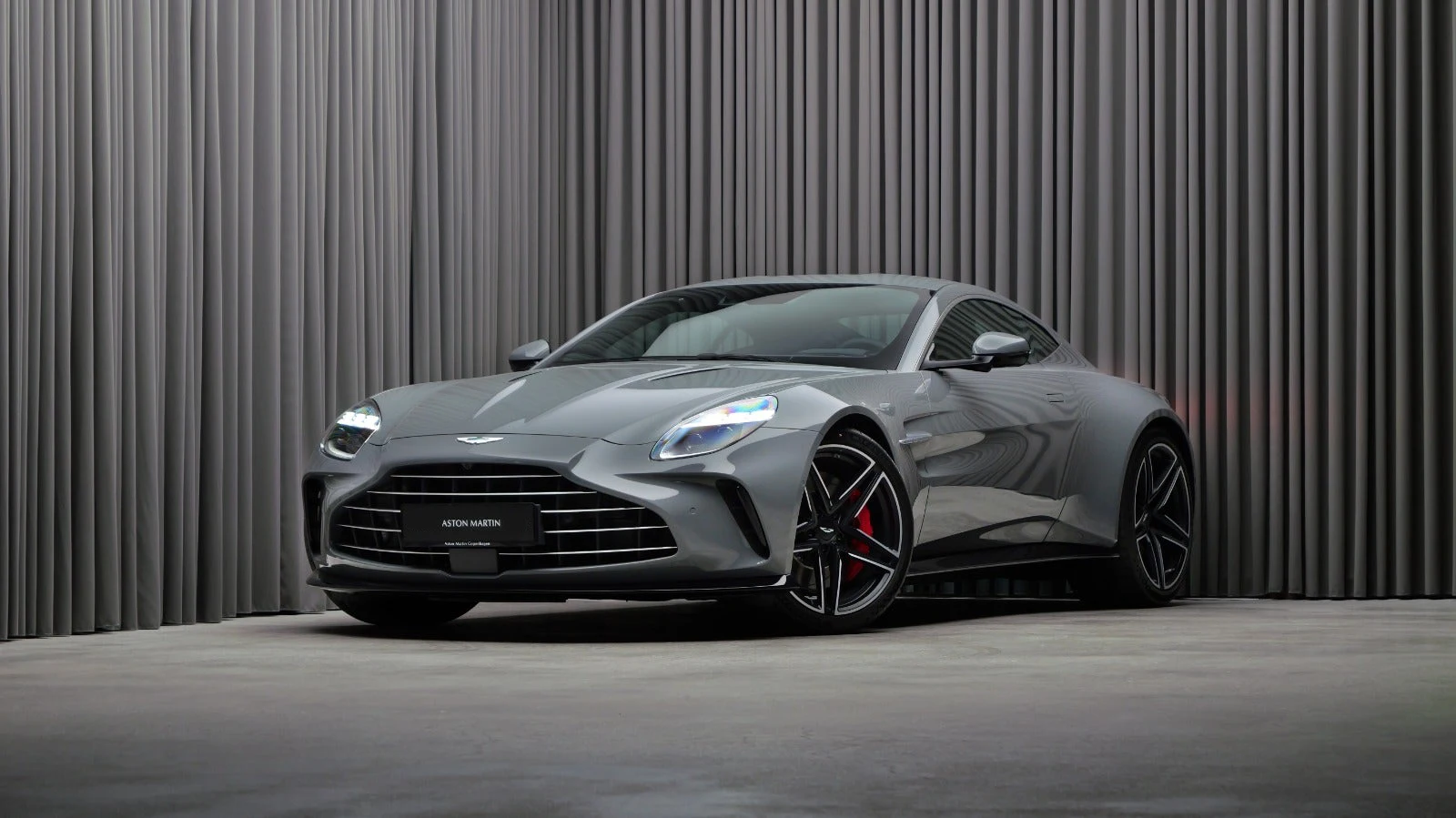 A grey Aston Martin Vantage standing in a showroom in front of a floor-to-ceiling pleated curtain, three-quarter front view, black diamond-cut wheels and red brake calipers