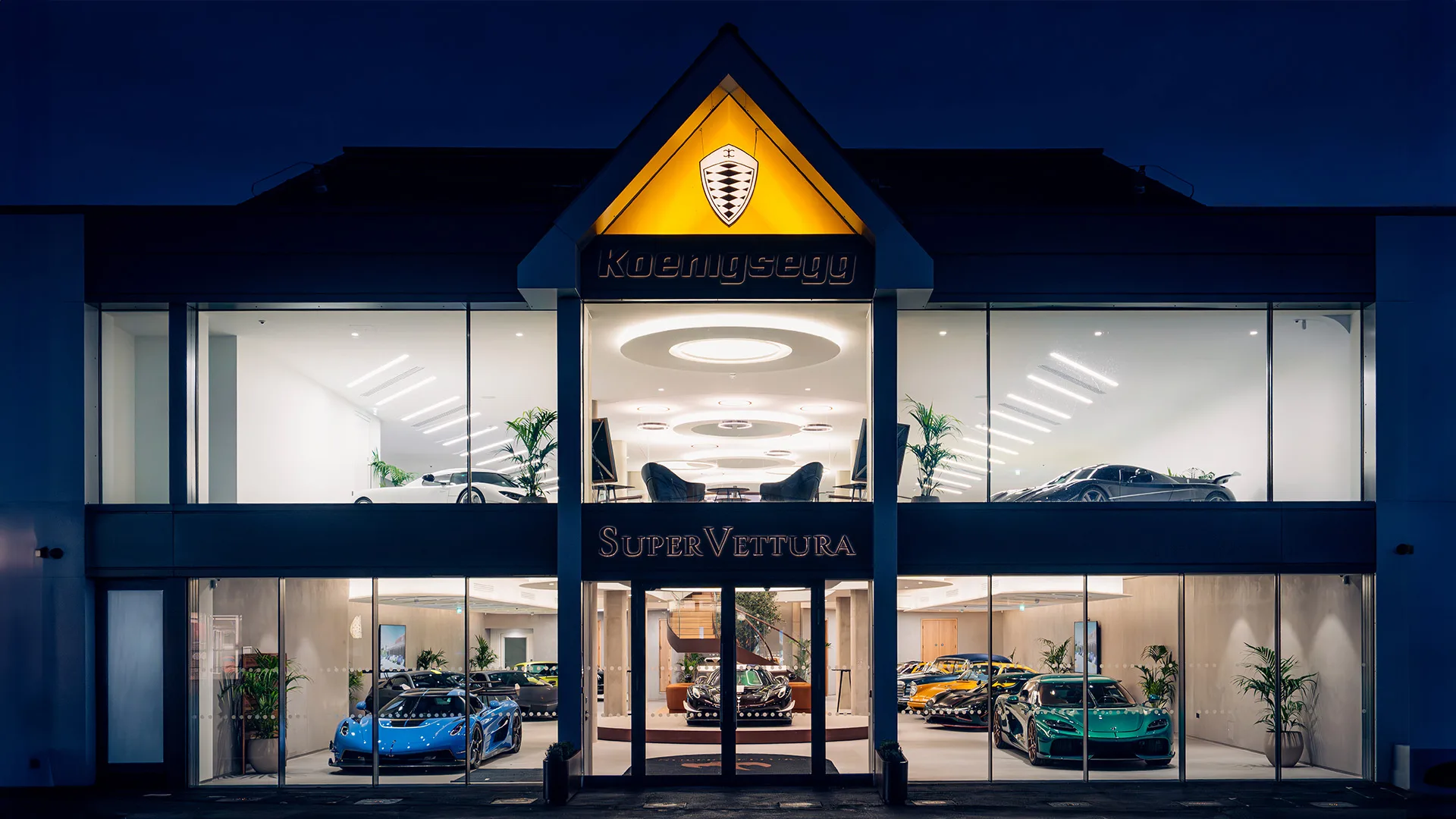 La façade du showroom SuperVettura au crépuscule à Sunningdale, avec l'enseigne Koenigsegg au-dessus de l'entrée et des hypercars visibles sur les deux niveaux